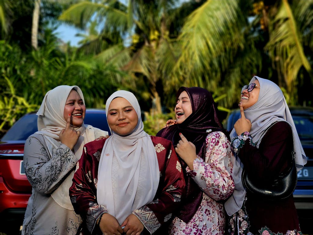 Four joyful Muslim women wearing hijabs enjoying the outdoors, surrounded by nature and parked cars.